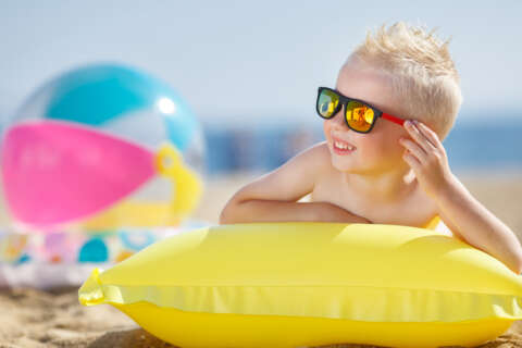 Krank im Urlaub: Ein kleiner Junge mit blonden kurzen Haaren trägt eine Sonnenbrille und liegt einer Luftmatratze lächelnd am Strand