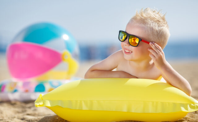 Krank im Urlaub: Ein kleiner Junge mit blonden kurzen Haaren trägt eine Sonnenbrille und liegt einer Luftmatratze lächelnd am Strand