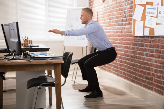 Bürosport Challenge: Ein Mann im Anzug macht hinter seinem Schreibtisch Kniebeugen