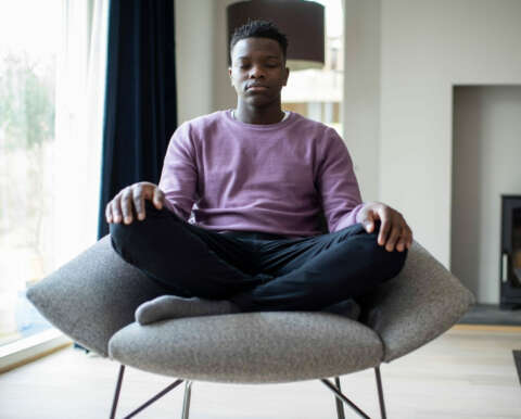 Meditation: Ein junger Mann sitzt im auf einem Sessel im Wohnzimmer und meditiert