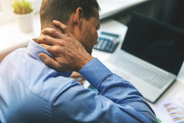 Sitzen: Ein Mann sitzt am Schreibtisch und massiert den Nacken w&auml;hrend er am Computer arbeitet