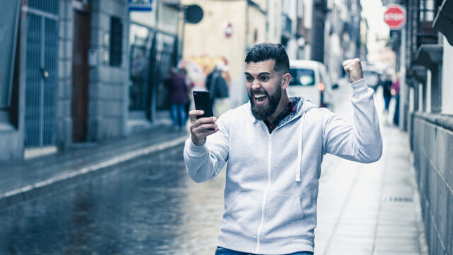 WM-Apps für den Fußball-Sommer: Junger Mann mit Bart schaut auf der Straße auf sein Smartphone und jubelt.