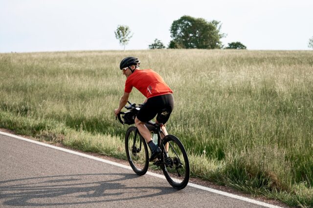 Ein Mann in rotem Radtrikot fährt mit seinem Rennrad auf einer Landstraße an Wiesen vorbei