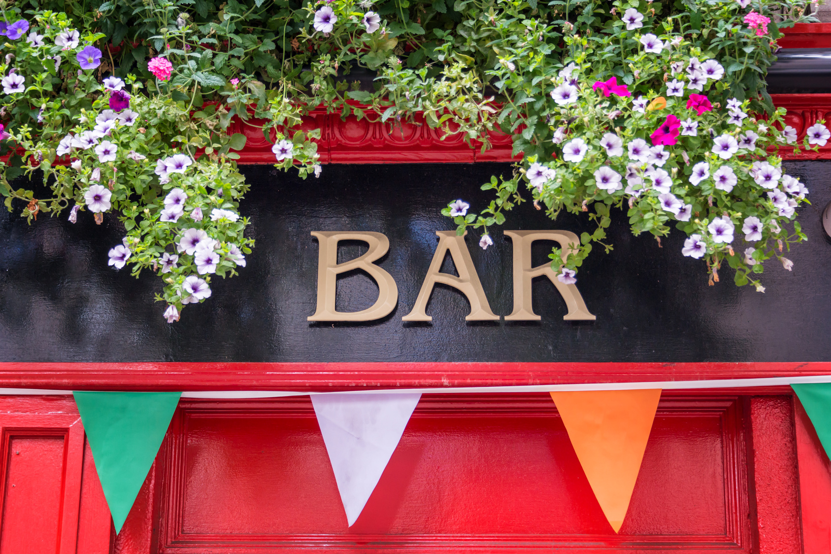 Das Wort Bar in Gold auf schwarzem Hintergrund über einer roten Holztür, vor der Wimpel in grün, weiß und orange hängen.