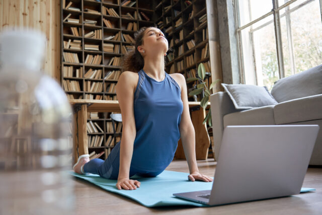 Junge Frau wiederholt Pilates-Übungen, die auf einem Laptop gezeigt werden.