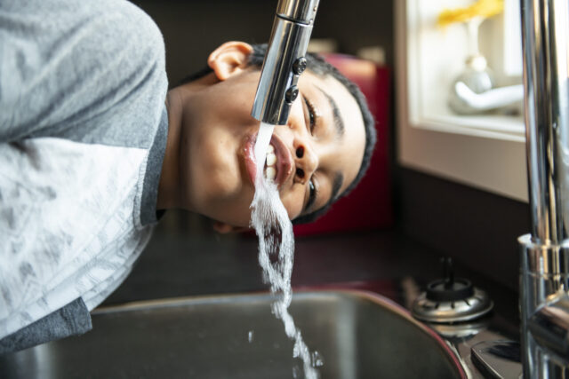 Wasser trinken: Ein Junge beugt sich in der K&uuml;che &uuml;ber das Sp&uuml;lbecken und trinkt aus dem Wasserkrahn