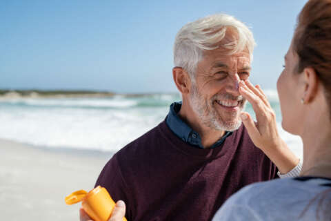 Sonnenschutz: Ein Mann steht im Pullover am Strand und wird auf der Nase mit Sonnencreme von einer Frau eingerieben.