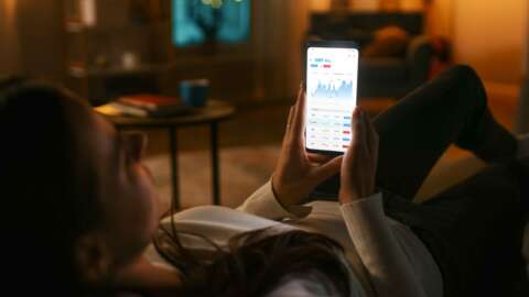 Spar dich glücklich: junge Frau liegt auf Sofa und schaut auf Smartphone um Börsenkurse zu prüfen.