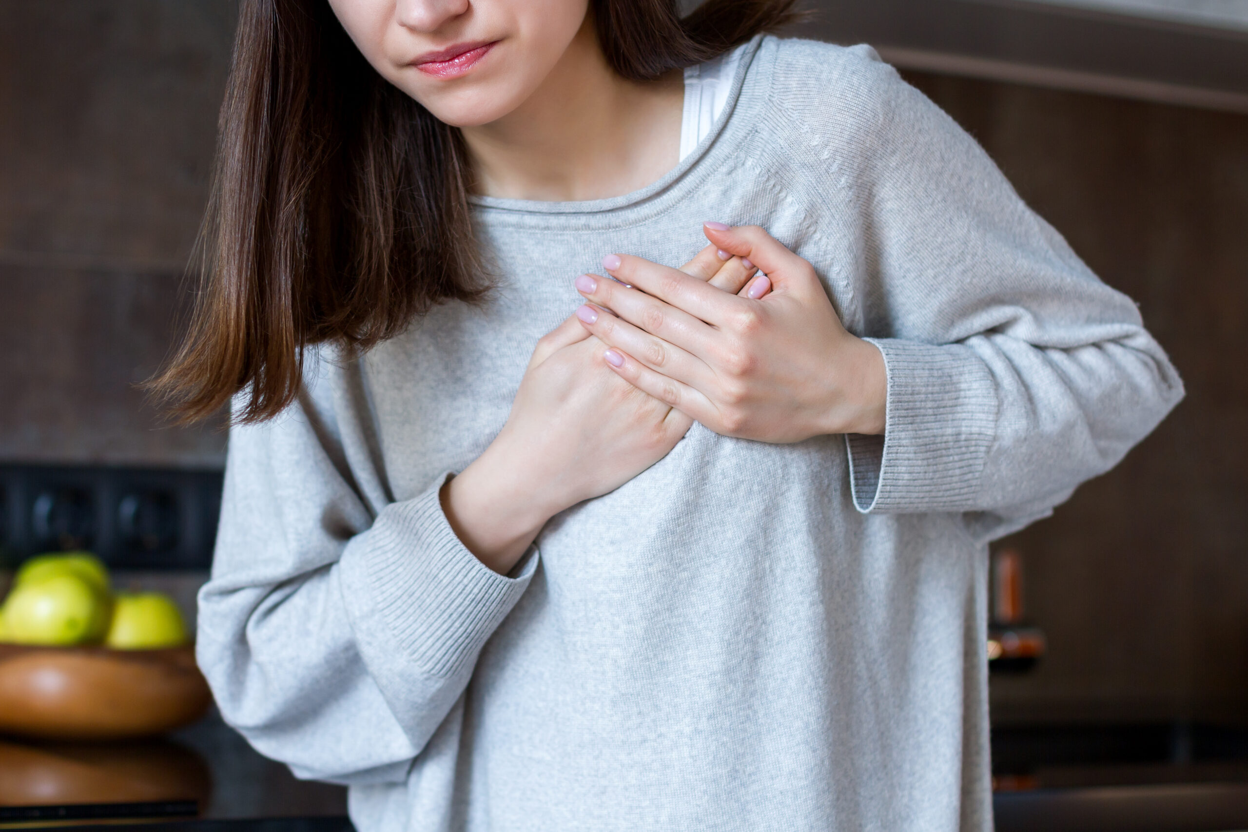 Organspende: Junge Frau mit grauem Pullover hält beide Hände an ihr Herz und scheint Schmerzen zu haben.