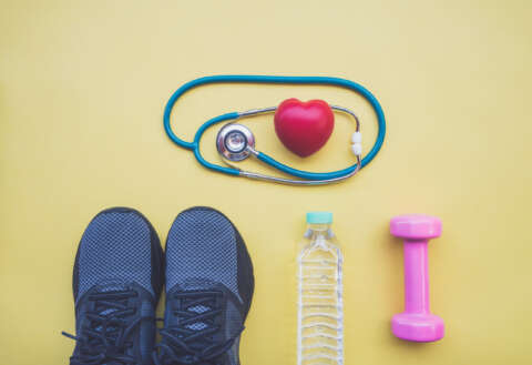 Sport nach Corona: Stethoskop mit Herz, Sportschuhe rosa Hantel und Wasserflasche auf gelbem Grund.