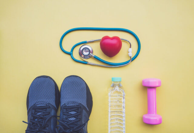 Sport nach Corona: Stethoskop mit Herz, Sportschuhe rosa Hantel und Wasserflasche auf gelbem Grund.
