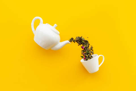 Tee: Weiße Teekanne und Tasse mit trockenen Teeblättern auf gelbem Grund.