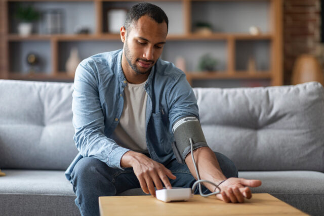 Big 5 Herzgesundheit: Young Black Man Sitting On Couch And Checking Blood Pressure With Upper Arm Monitor, Millennial African American Guy Measuring Arterial Tension While Relaxing In Living Room At Home, Copy Space